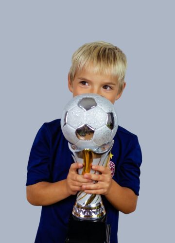 Young soccer player in blue jersey holds winners cup after the goal, isolated at light background